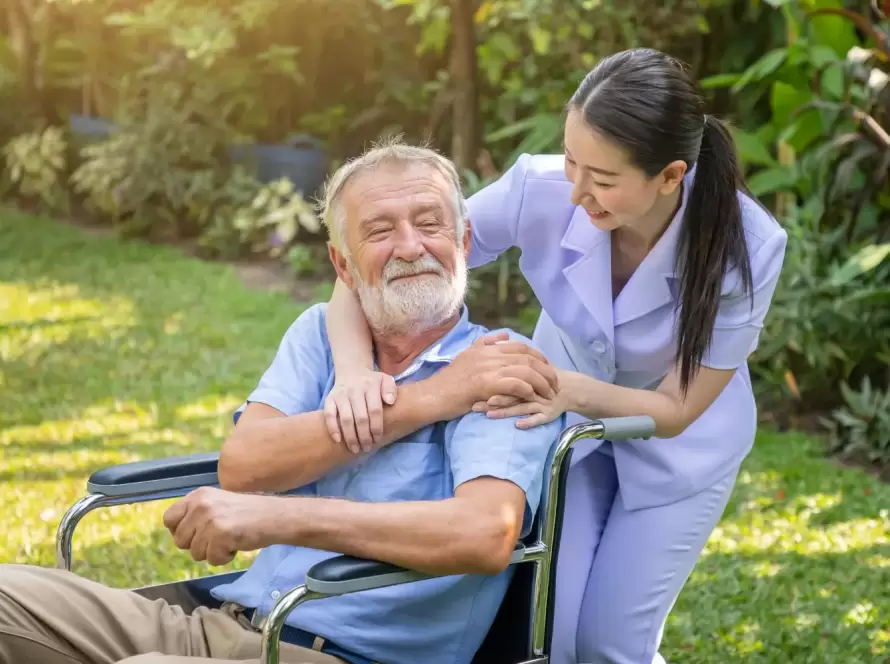 Anciano sonriente en silla de ruedas, recibiendo atención y compañía de una cuidadora en un jardín. La escena refleja calidez y cuidado en el cuidado de personas mayores.
