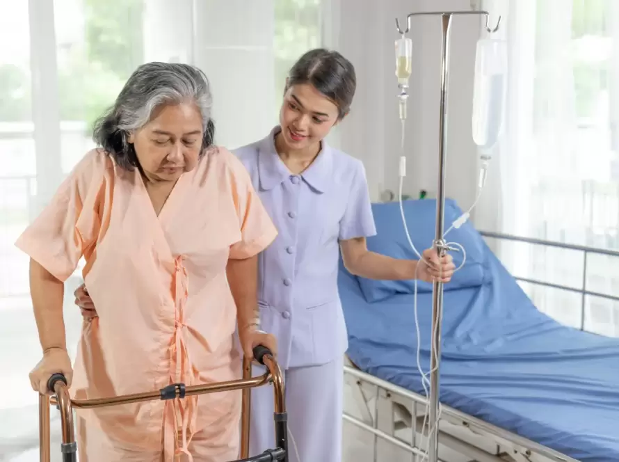 Anciana en un hospital recibiendo asistencia de una enfermera mientras usa un andador, con un gotero en el fondo y un ambiente luminoso y acogedor.