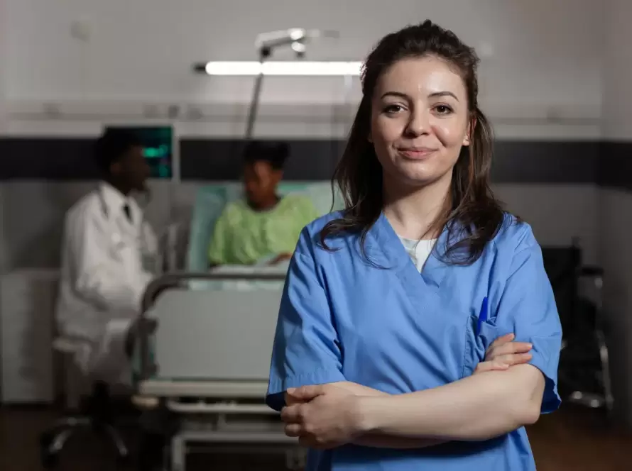 Mujer enfermera en uniforme azul sonriente, de pie en un hospital, con pacientes en el fondo recibiendo atención médica. La imagen destaca la dedicación y profesionalismo en el ámbito de la salud.